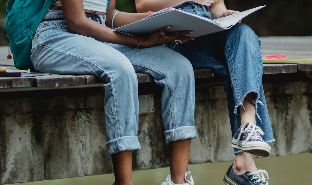 Crop unrecognizable multiracial female students in jeans with backpack reading new material in textbook together while sitting on wooden pier in green summer park